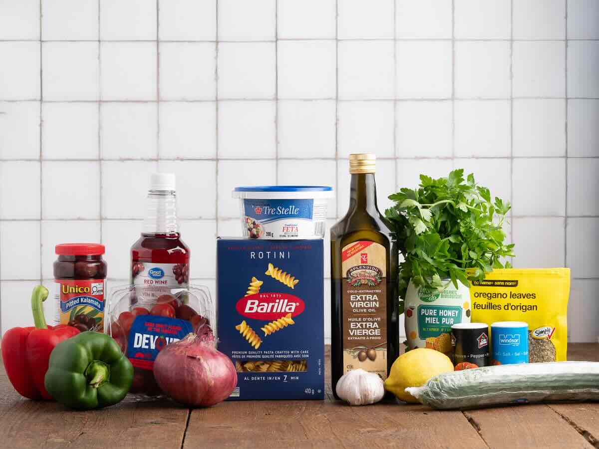 A variety of ingredients on a wooden table, perfect for creating a delicious Greek Pasta Salad, including pasta, vegetables, olive oil, red wine vinegar, feta cheese, olives, parsley, lemon, garlic, oregano, and honey—all beautifully arranged in front of a white tiled wall.