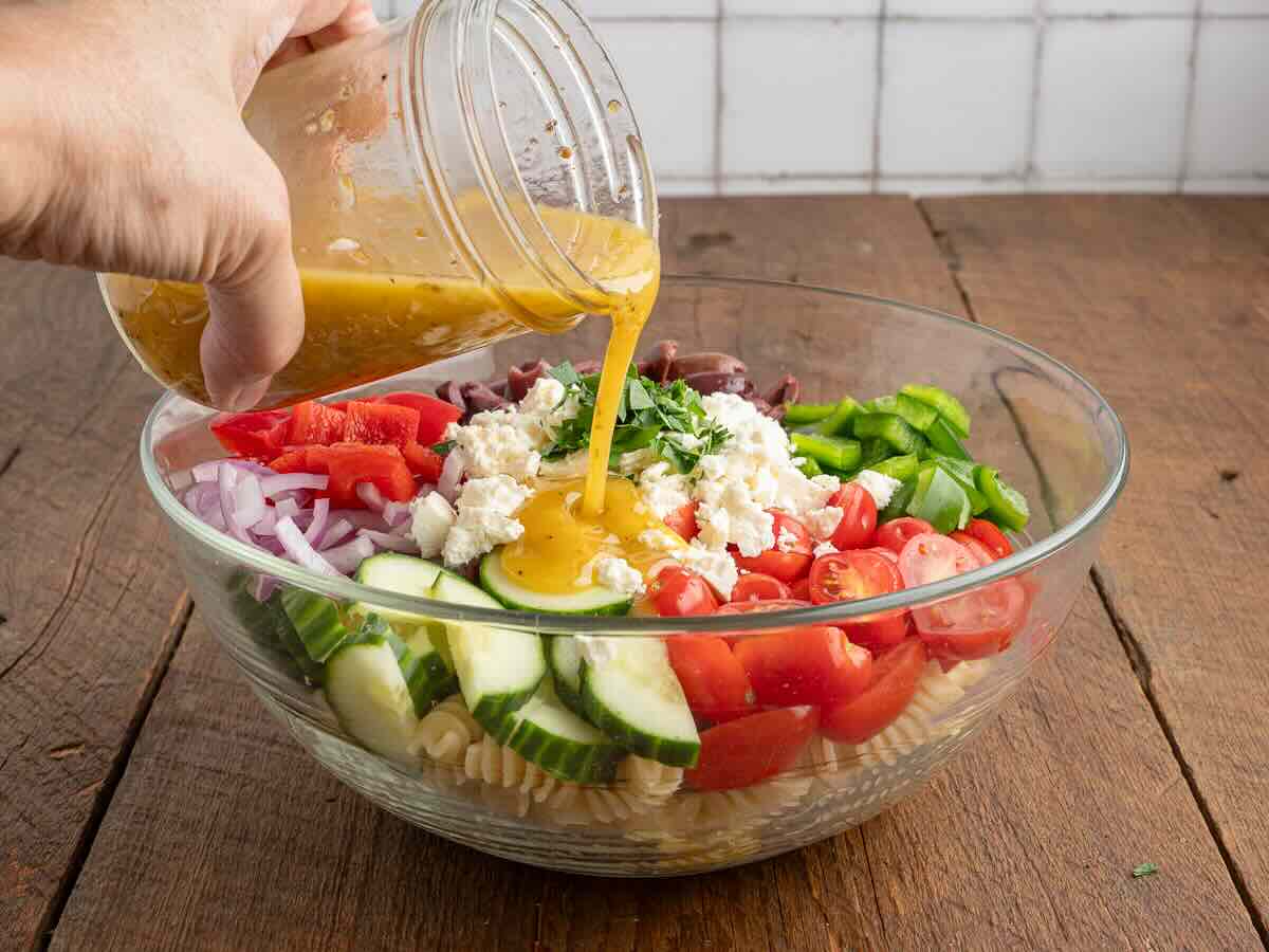 A hand pours dressing from a jar onto a glass bowl filled with pasta, cucumbers, tomatoes, onions, peppers, feta cheese, and herbs—all the delicious ingredients of a classic Greek Pasta Salad—set on a wooden table.