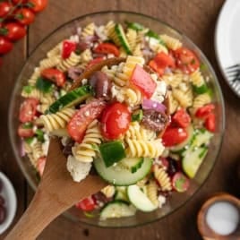 A wooden spoon scoops a serving of Greek Pasta Salad with tomatoes, cucumber, olives, red onion, and feta from a glass bowl—a vibrant twist inspired by a classic Greek Salad Recipe.