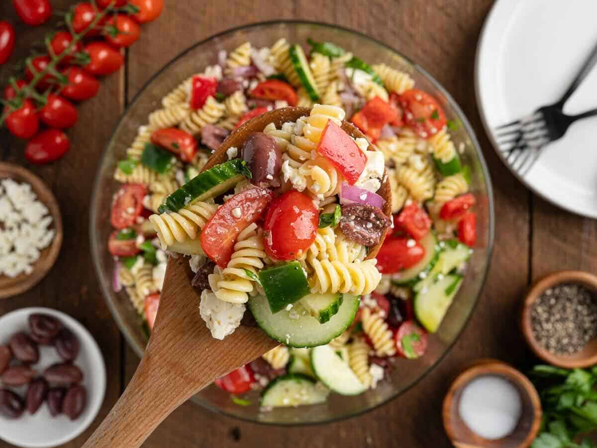 A wooden spoon scoops a serving of Greek Pasta Salad with tomatoes, cucumber, olives, red onion, and feta from a glass bowl—a vibrant twist inspired by a classic Greek Salad Recipe.