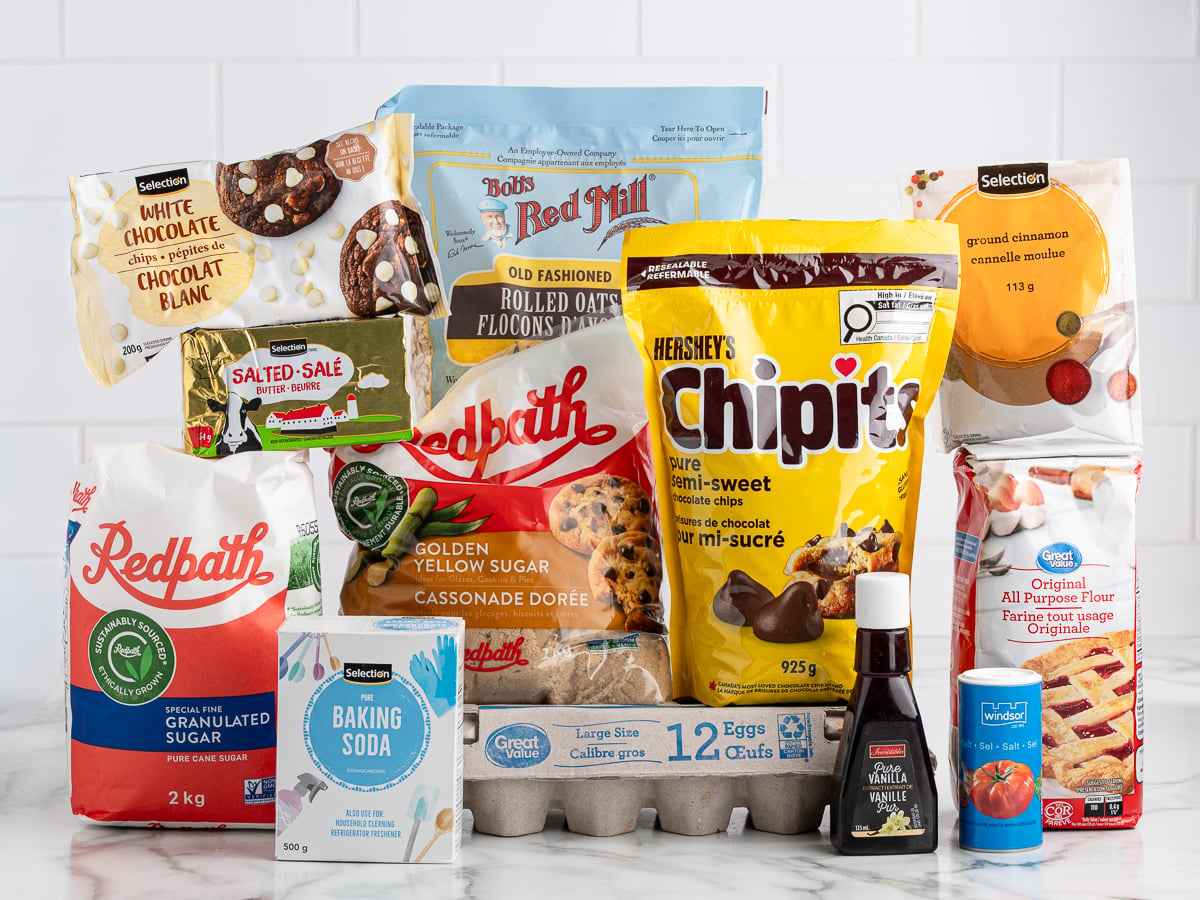 A selection of baking ingredients on a white surface, perfect for making Marry Me Cookies&mdash;sugar, brown sugar, flour, eggs, chocolate chips, white chocolate, oats, baking soda, vanilla extract, cinnamon, and salt&mdash;all in their original packaging.