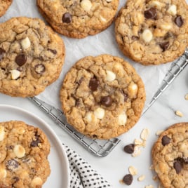 Several freshly baked Marry Me Cookies with white and dark chocolate chips are cooling on a wire rack and plate, with oats and chocolate pieces scattered nearby on a light surface.