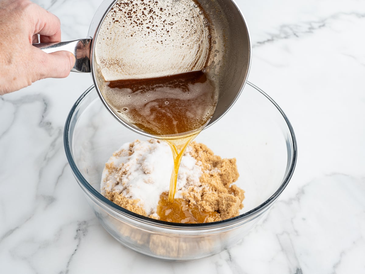 A hand pours melted butter from a saucepan into a glass bowl containing brown sugar and granulated sugar on a white marble countertop, beginning the delicious base for Marry Me Cookies.
