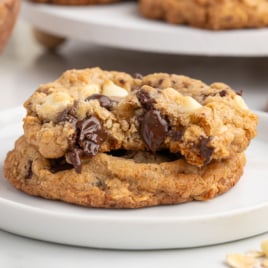 Two Marry Me Cookies stacked on a white plate, featuring gooey melted chocolate and white chocolate chips, with more cookies blurred in the background.