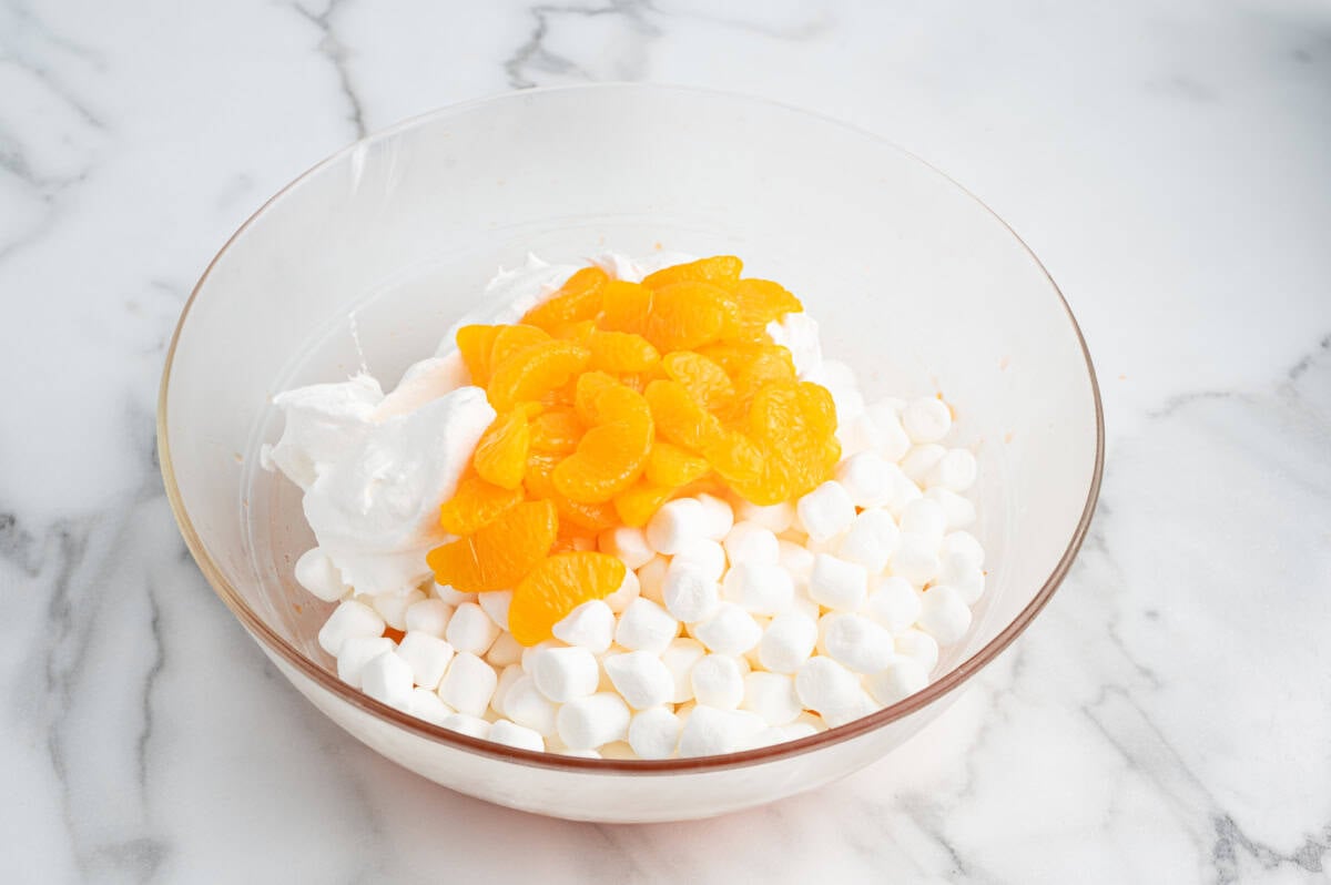 A clear glass bowl on a marble surface contains mini marshmallows, mandarin orange segments, and a scoop of whipped topping—classic ingredients for an easy Orange Creamsicle Salad.