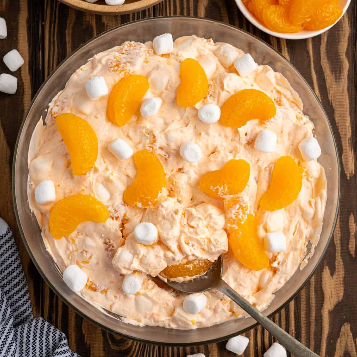 A glass bowl filled with creamy Orange Creamsicle Salad, decorated with canned mandarin orange segments and mini marshmallows. A spoon rests in the bowl, ready to serve this delightful Orange Creamsicle Salad Recipe.