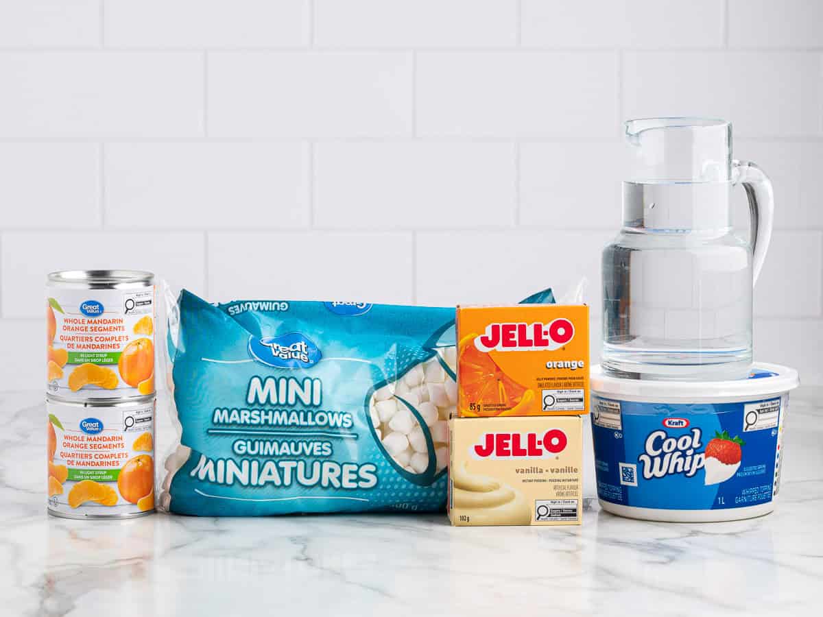 Various ingredients for an Orange Creamsicle Salad are on a marble counter, including canned mandarin oranges, mini marshmallows, orange and vanilla Jello boxes, a pitcher of water, and Cool Whip, set against a white tiled wall.