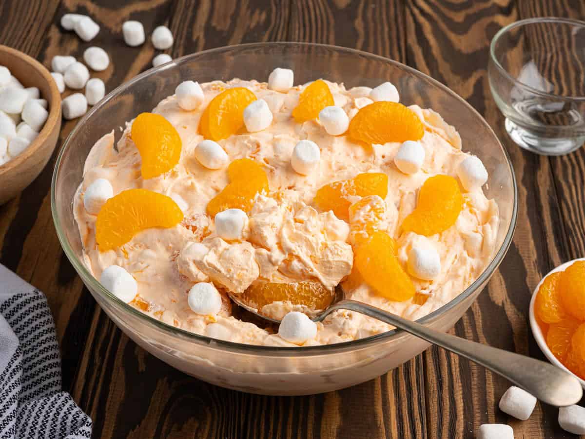 A glass bowl filled with Orange Creamsicle Salad, topped with mandarin segments and mini marshmallows, sits with a spoon inside and extra marshmallows and oranges nearby on a wooden table.