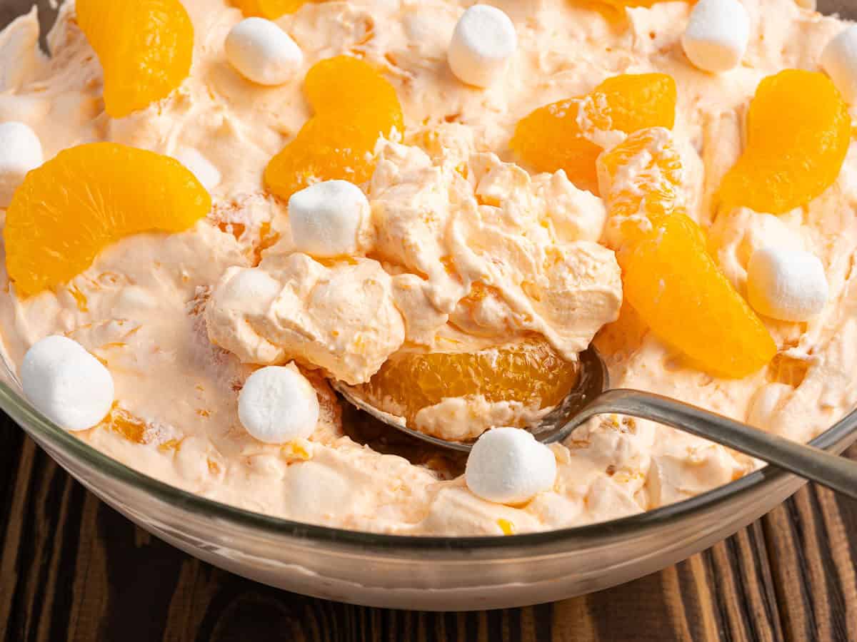A bowl of creamy Orange Creamsicle Salad topped with mandarin orange slices and mini marshmallows. A spoon scoops up the fluffy mix, revealing the delicious blend of ingredients. The bowl rests on a wooden surface.