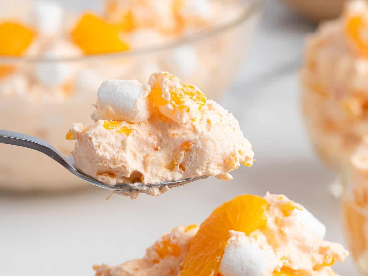 A close-up of a spoonful of creamy Orange Creamsicle Salad with mini marshmallows and mandarin orange slices, featuring the vibrant dessert in focus as the bowl of this Orange Creamsicle Salad Recipe sits blurred in the background.