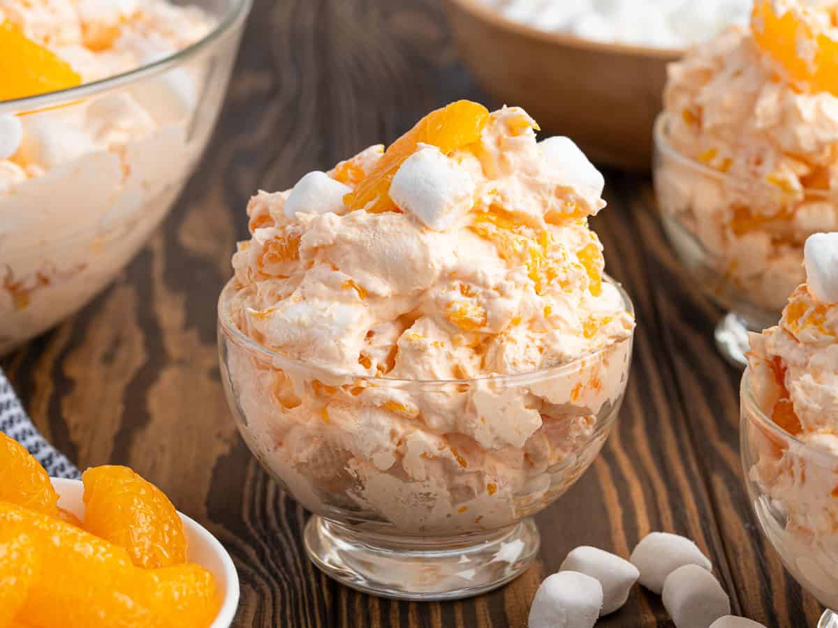 A glass bowl filled with creamy Orange Creamsicle Salad, topped with mandarin orange slices and mini marshmallows, sits on a wooden surface with more dessert bowls and ingredients nearby.