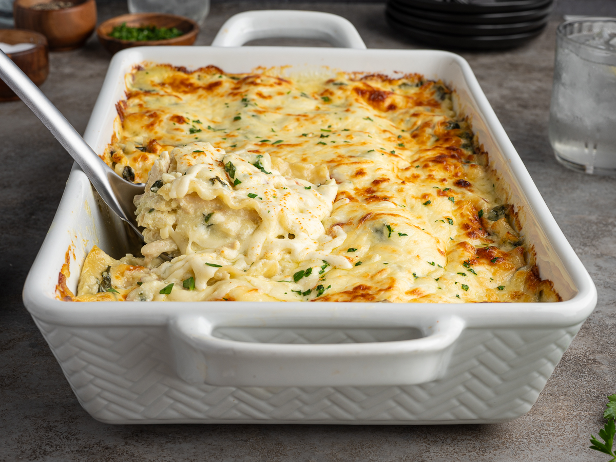A baked cheesy white lasagna with cream cheese in a white casserole dish, with a serving spoon lifting out a portion. The lasagna is golden brown on top and garnished with chopped herbs.
