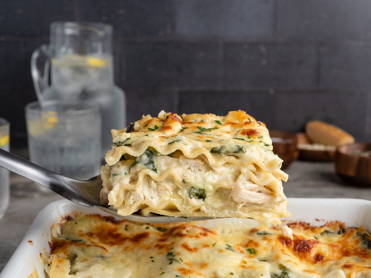 A close-up of a spatula lifting a cheesy, golden-brown slice of white chicken lasagna recipe from a baking dish. In the background, there are glasses and a pitcher of water with lemon slices.