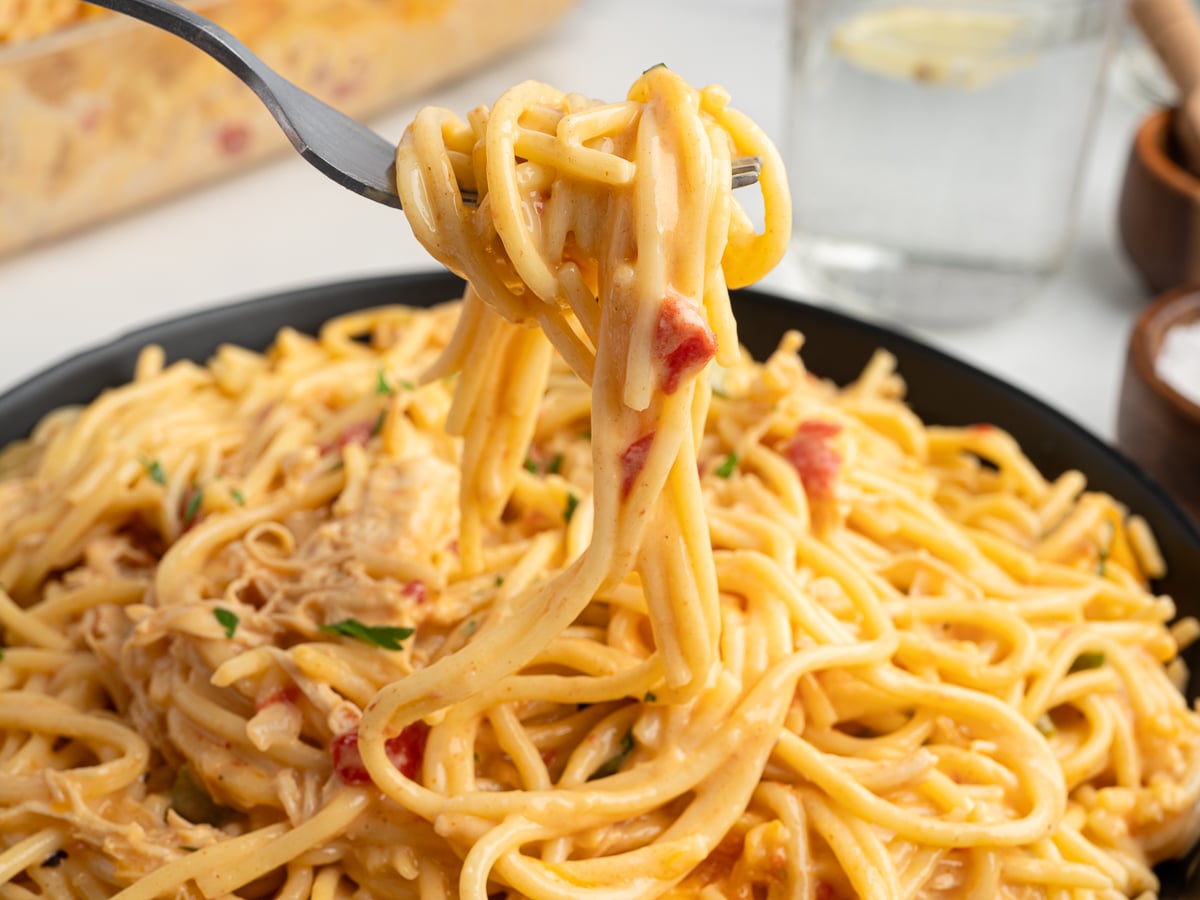 A fork lifts a serving of creamy Rotel Chicken Spaghetti with pieces of chicken and diced tomatoes from a black bowl. A glass of water with lemon and small bowls are visible in the background.