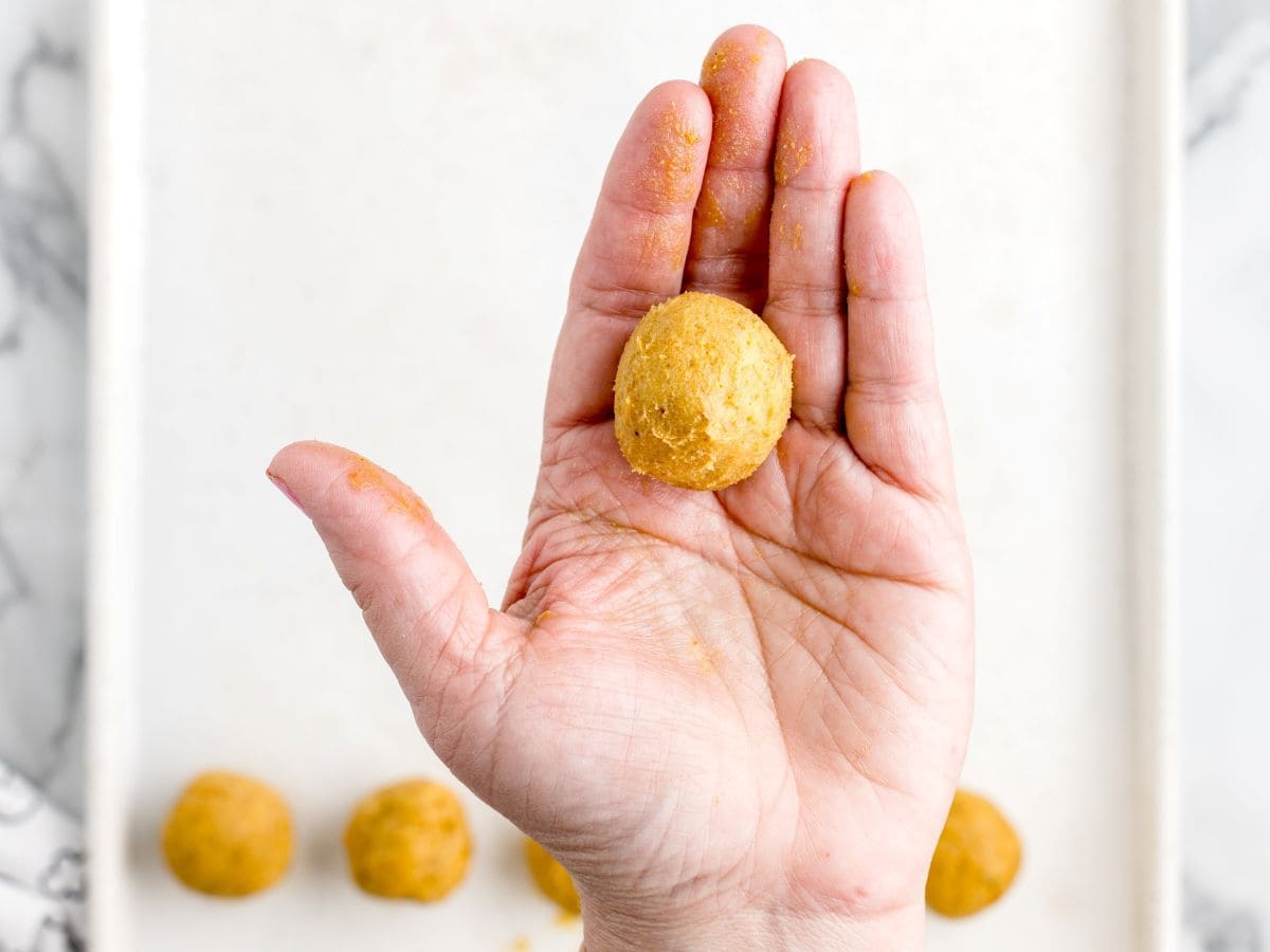 A hand holds a round ball of dough, resembling delicious no bake cheesecake balls, with more dough balls resting on a white surface in the background.