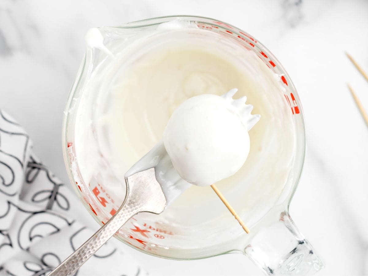 A cake pop on a stick is being coated in white chocolate using a fork, held above a glass measuring cup filled with melted white chocolateโperfect for making no bake cheesecake balls or pumpkin cheesecake treats.