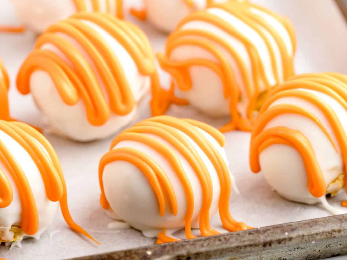 White chocolate truffles with orange icing drizzle are arranged on a parchment-lined baking sheet, ready to be servedโthese no bake cheesecake balls offer a delightful twist on classic treats.
