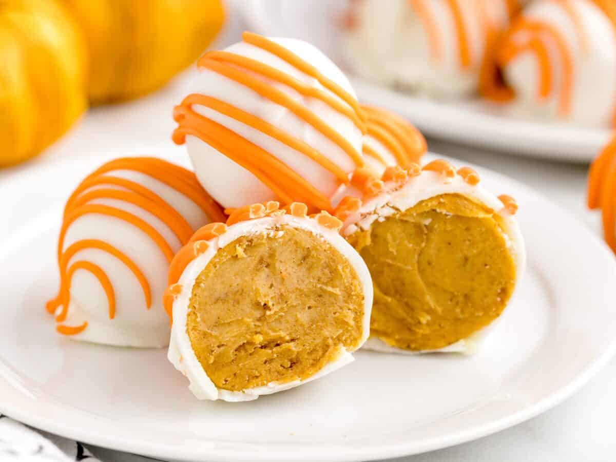 A plate of round pumpkin cheesecake truffles, also known as no bake cheesecake balls, are coated in white chocolate with orange drizzle on top. One truffle is cut in half, revealing the creamy pumpkin cheesecake filling inside. Pumpkins are blurred in the background.