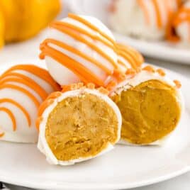 A plate of round pumpkin cheesecake truffles, also known as no bake cheesecake balls, are coated in white chocolate with orange drizzle on top. One truffle is cut in half, revealing the creamy pumpkin cheesecake filling inside. Pumpkins are blurred in the background.