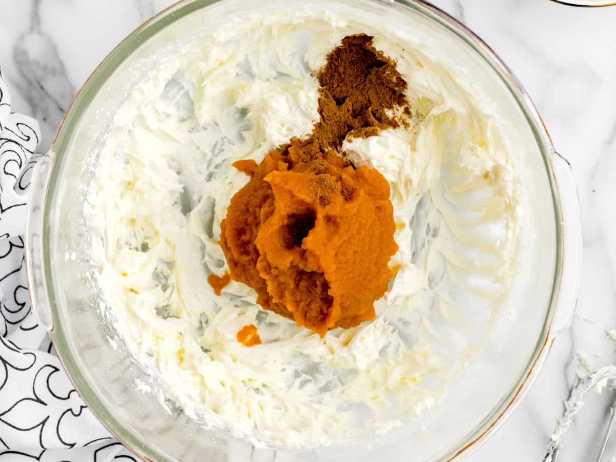 A glass mixing bowl with cream cheese, pumpkin puree, and spices sits on a marble countertop, ready to be mixed for a delicious batch of no bake pumpkin cheesecake balls.