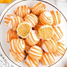 A plate of round white chocolate cheesecake balls drizzled with orange icing, with two truffles cut in half to reveal a creamy orange filling inside; these no bake treats are the perfect twist on classic pumpkin cheesecake.