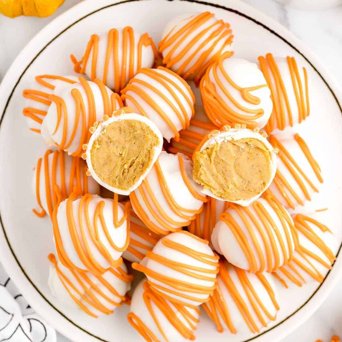 A plate of round white chocolate cheesecake balls drizzled with orange icing, with two truffles cut in half to reveal a creamy orange filling inside; these no bake treats are the perfect twist on classic pumpkin cheesecake.