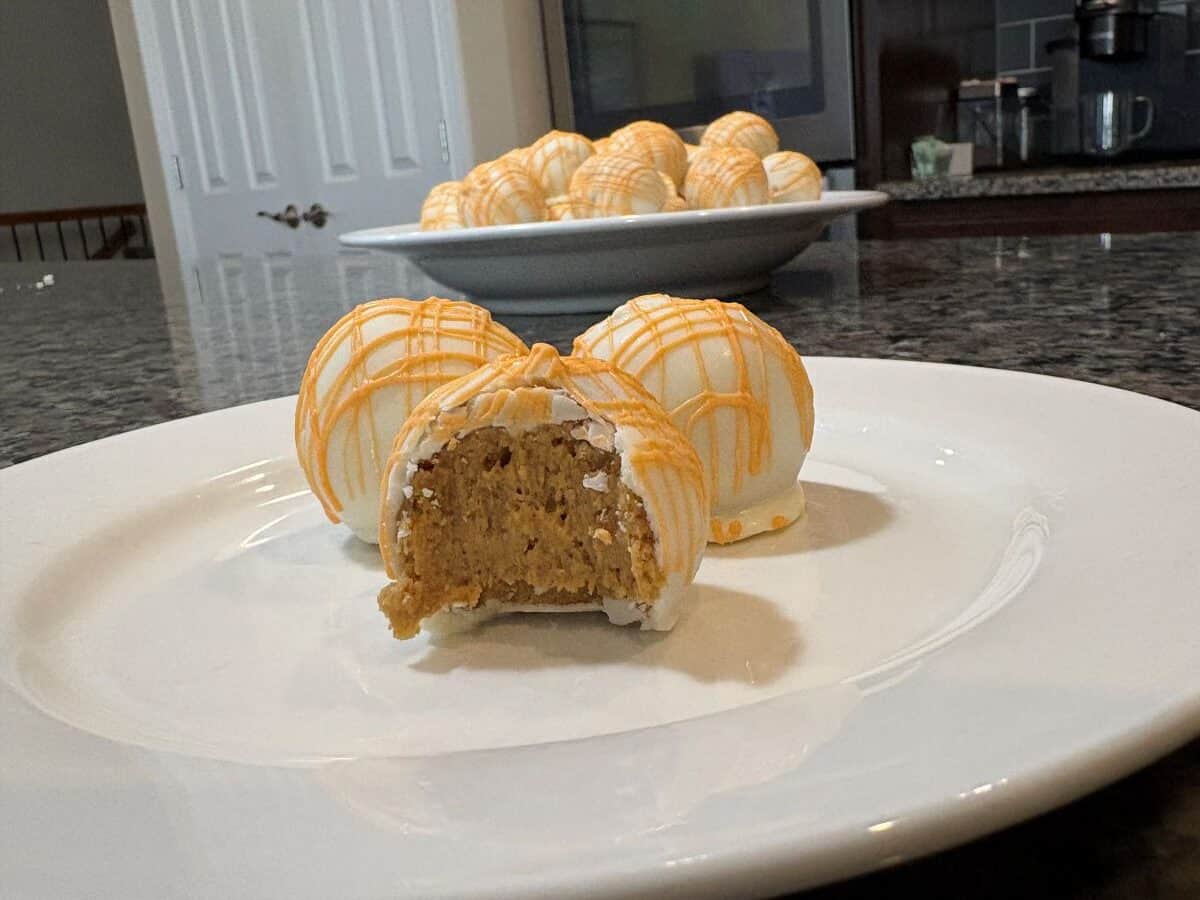 A close-up of white chocolate-coated cheesecake balls with orange drizzle on a plate, one with a bite taken out, and more no bake cake balls on a platter in the background on a granite countertop.