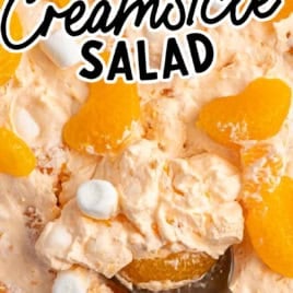 A bowl of creamy Orange Creamsicle Salad topped with mandarin orange slices and mini marshmallows. A spoon is scooping some of the salad. Text overlay reads, The Best Orange Creamsicle Salad Recipe: Vintage Recipe!.