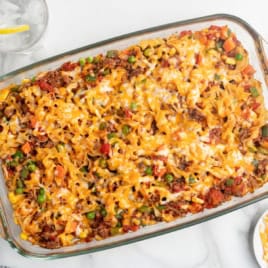 A glass baking dish filled with a cheesy noodle casserole, this classic Hamburger Casserole is topped with melted cheese, ground meat, peas, carrots, and diced tomatoes. It sits on a white surface next to a plate and a glass of water with a lemon slice.