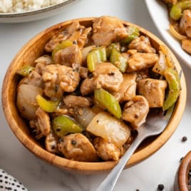 A wooden bowl filled with stir-fried chicken pieces, sliced celery, and onions in a savory Panda Express Pepper Chicken-style sauce, with a fork resting inside. A plate of rice is partially visible in the background.
