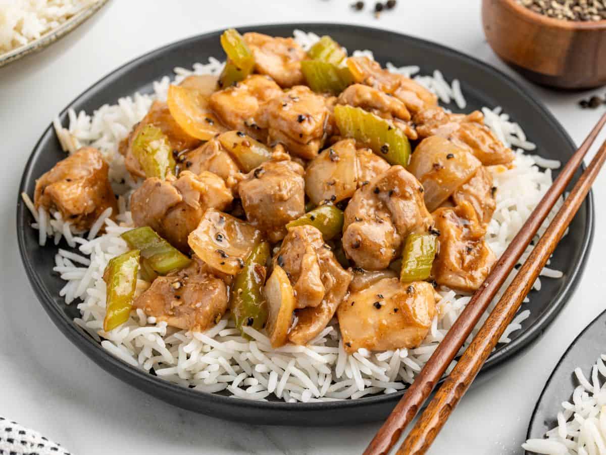 A black plate of white rice topped with Panda Express Pepper Chickenโtender pieces of chicken, celery, and onions in a savory brown sauce, garnished with black pepper. Wooden chopsticks rest on the side of the plate.