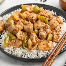 A black plate filled with white rice topped with chunks of chicken, celery, and onions in a savory brown sauce, reminiscent of Panda Express Pepper Chicken. Chopsticks rest on the edge of the plate.