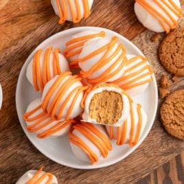A plate of round white truffles with orange drizzle, one with a bite showing a light brown filling, surrounded by cookies and a small bowl of spice on a wooden surface—these no bake Pumpkin Cheesecake Balls are the perfect fall treat.