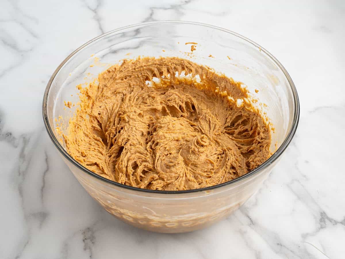 A glass bowl filled with creamy, light brown cookie dough sits on a white marble surface. The dough looks freshly mixed, with swirls and peaks visible—perfect for making no bake Pumpkin Cheesecake Balls that capture the flavors of fall in every bite.