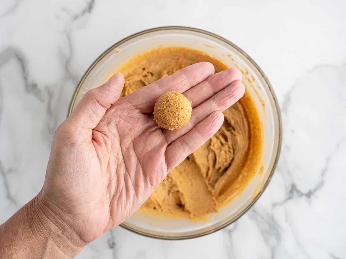 A hand holds a small, round ball of no bake cheesecake dough above a glass bowl filled with more dough on a white marble surface.