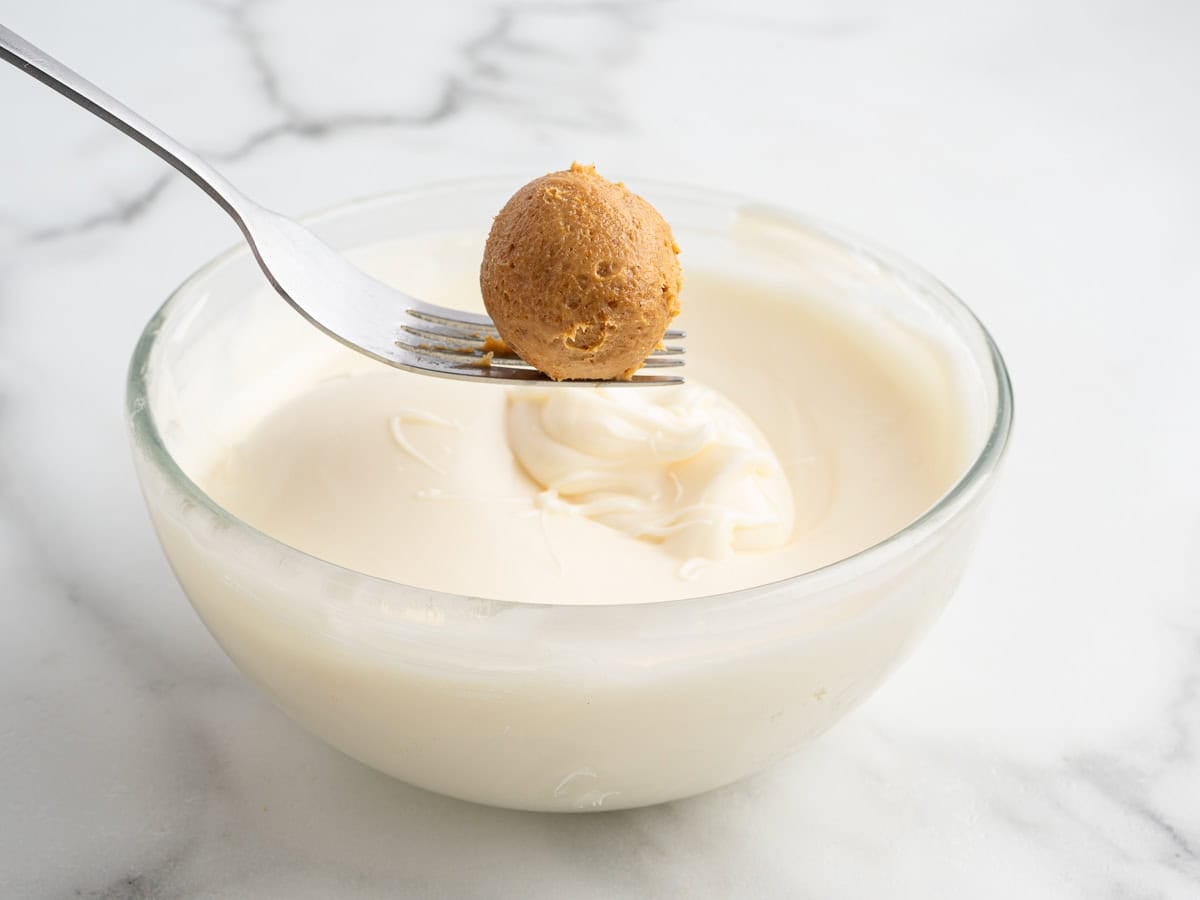 A fork holds a round Pumpkin Cheesecake Ball of dough above a glass bowl filled with melted white chocolate on a marble surface, perfect for making these irresistible no bake treats.
