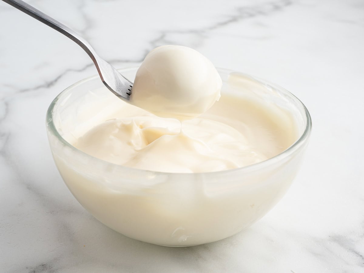 A clear glass bowl filled with thick, creamy white yogurt sits on a marble surface, reminiscent of the rich texture of no bake pumpkin cheesecake. A spoon is lifting a dollop of the yogurt from the bowl, inviting you to imagine transforming it into delectable cheesecake balls.