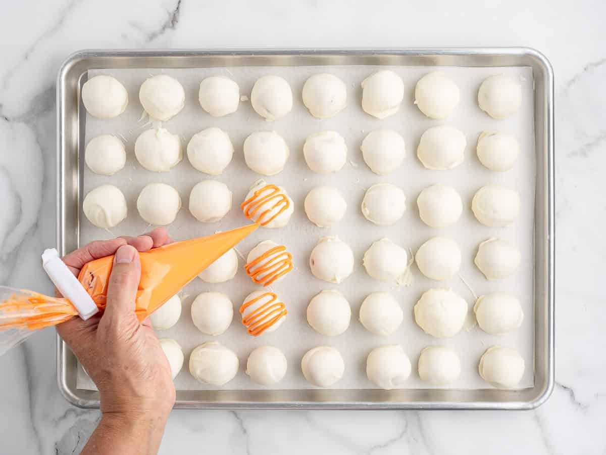 A hand uses an orange piping bag to drizzle icing on No Bake Pumpkin Cheesecake Balls covered in white chocolate, arranged neatly on a parchment-lined baking sheet.