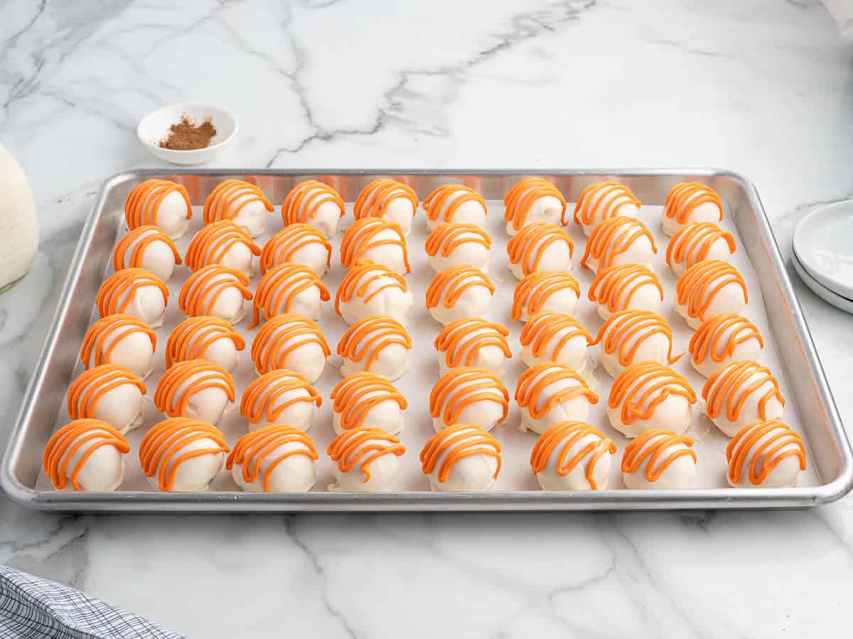 A baking tray holds rows of round white Cheesecake Balls, topped with orange icing drizzle reminiscent of Pumpkin Cheesecake, set on a marble countertop. A small bowl of cinnamon and part of a towel are visible nearby, perfect for this easy no bake treat.