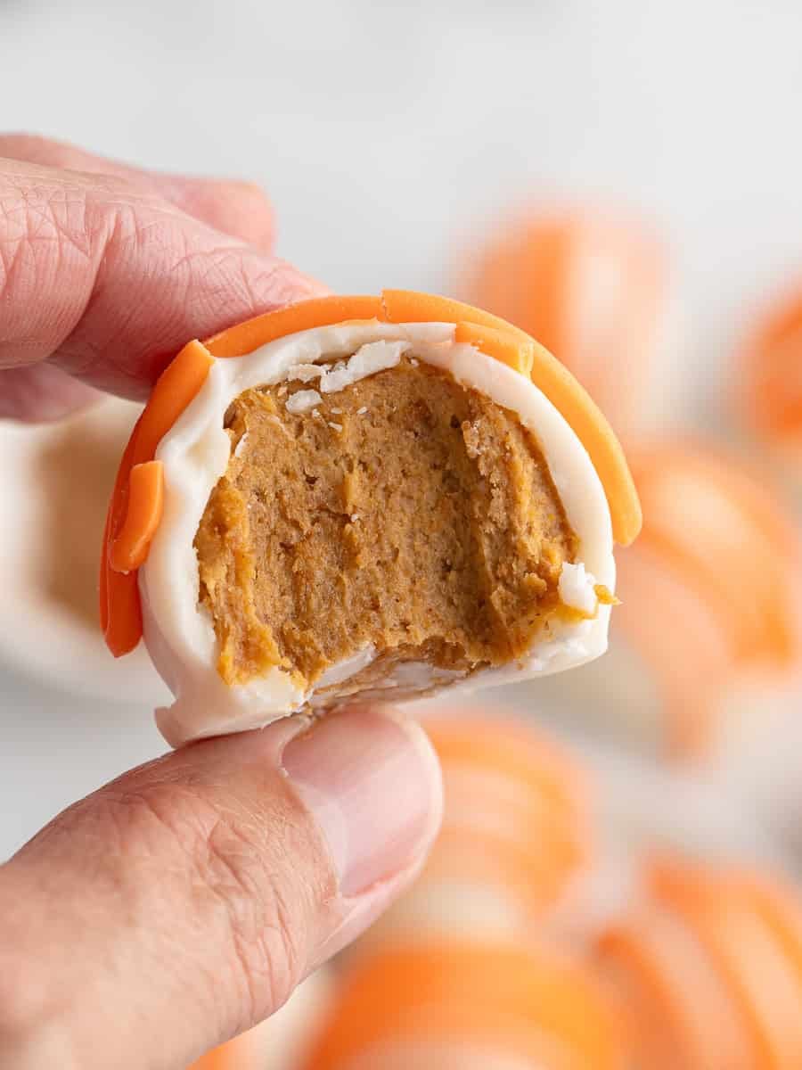 A hand holding a white chocolate-covered treat with an orange swirl, showing a creamy orange-brown filling with a bite taken out—these no bake Pumpkin Cheesecake Balls offer the perfect blend of rich cheesecake flavor and autumn-inspired sweetness.