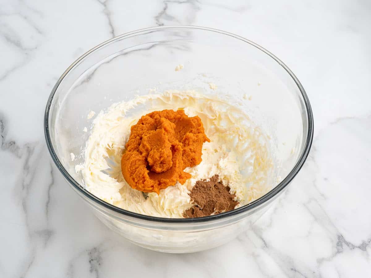 Clear glass bowl on a marble surface containing cream cheese, a mound of pumpkin puree, and a small pile of brown spices, ready to be mixed together for delicious No Bake Pumpkin Cheesecake Balls.