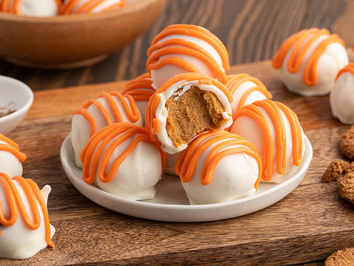A plate of white chocolate truffles with orange drizzle, one with a bite showing a soft, brown filling inside—these no bake cheesecake balls are irresistibly creamy. More truffles and cookies are in the background on a wooden surface, adding to the festive pumpkin cheesecake-inspired treat display.