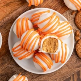 A plate of round white Cheesecake Balls with orange drizzle, one with a bite showing a creamy brown Pumpkin Cheesecake filling, on a wooden surface with cookies nearby—an irresistible no bake treat.