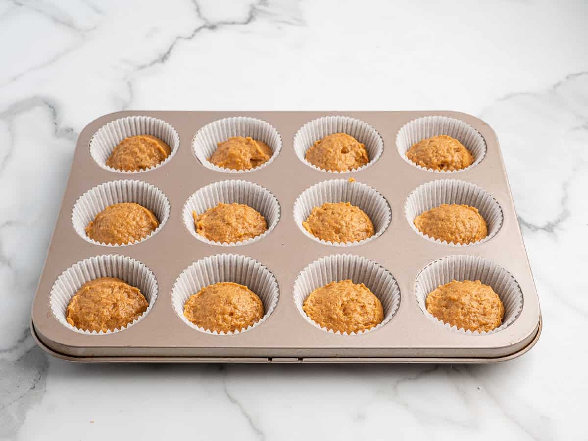 A muffin tin lined with white paper cups is filled with unbaked batter for Pumpkin Cream Cheese Muffins, sitting on a white marble surface.