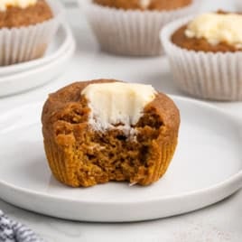 A Pumpkin Cream Cheese Muffin with a smooth cream cheese filling and a bite taken out sits on a white plate. Other whole Pumpkin Cream Cheese Muffins in white paper liners are visible in the background, making this Pumpkin Cream Cheese Muffins Recipe look especially inviting.