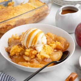 A bowl of apple cobbler topped with vanilla ice cream and caramel sauce, with a spoon. There are apples, caramel candies, a pitcher of caramel, and a dish of cobbler in the backgroundโcapturing the cozy essence of a Caramel Apple Dump Cake Recipe in every delicious detail.