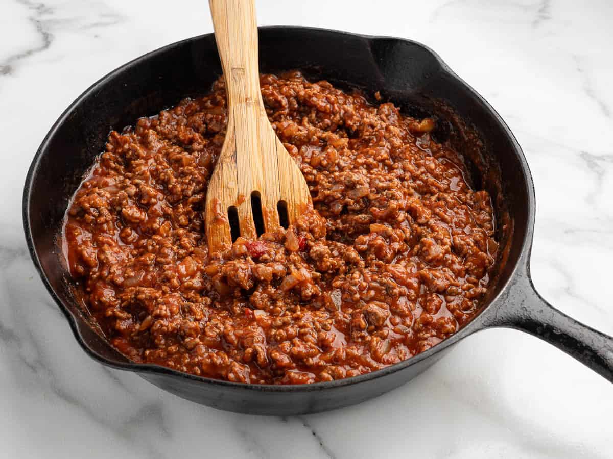A cast iron skillet filled with cooked ground beef in a rich tomato sauce, perfect for Garlic Bread Sloppy Joes, is stirred with a wooden spatula and sits on a white marble surface.