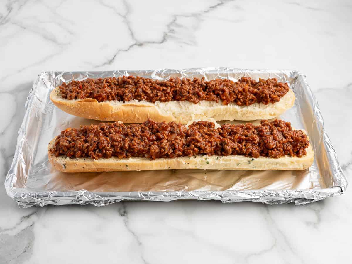 Two hoagie rolls filled with a savory ground meat mixture sit on a foil-lined baking sheet on a marble surface, ready to become delicious Garlic Bread Sloppy Joes.