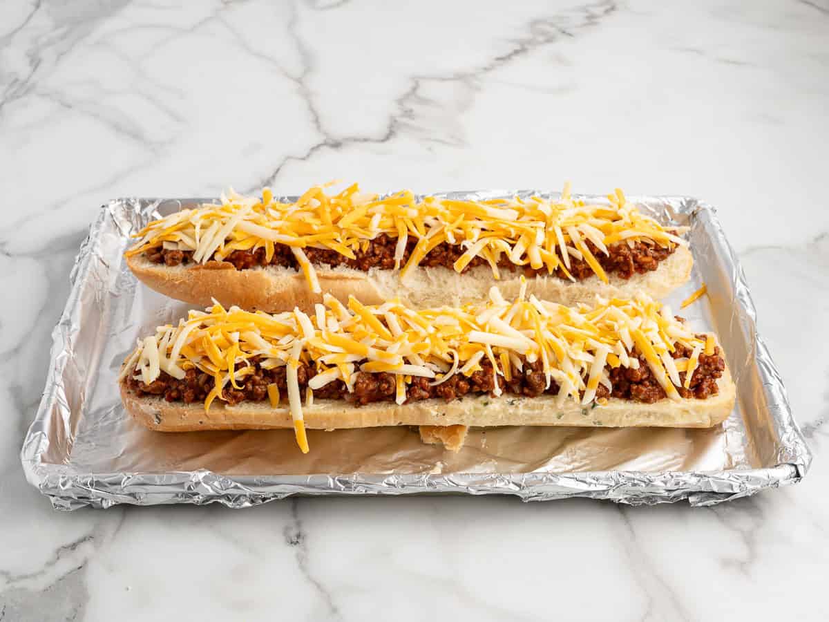 Two open-faced sandwich halves topped with cooked meat and shredded cheese rest on a foil-lined baking sheet on a marble countertop, creating a mouthwatering take on Garlic Bread Sloppy Joes.