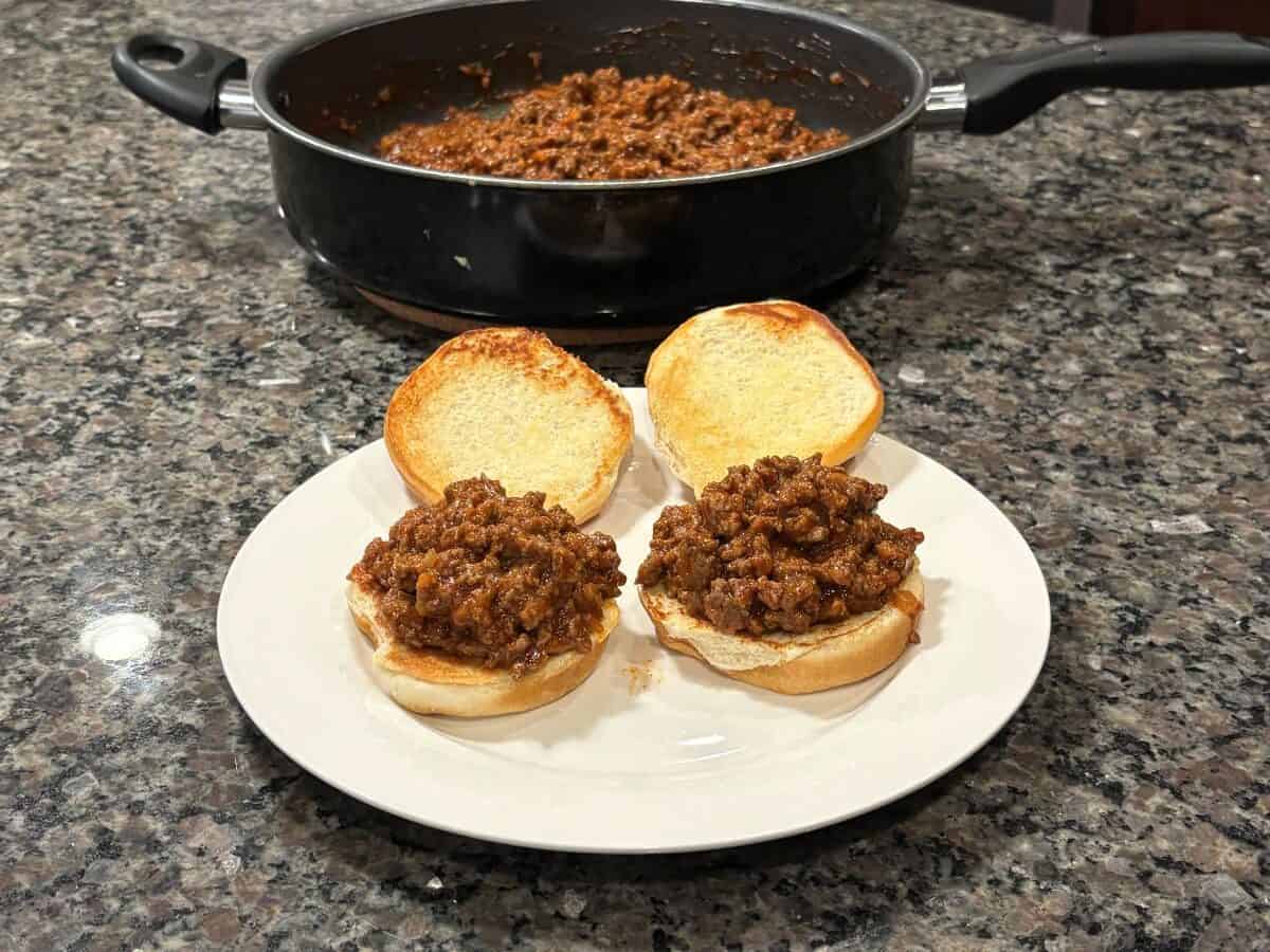 Two open Sloppy Joes sandwiches sit invitingly on a white plate atop a granite countertop, with a skillet full of hearty Sloppy Joes filling in the background.